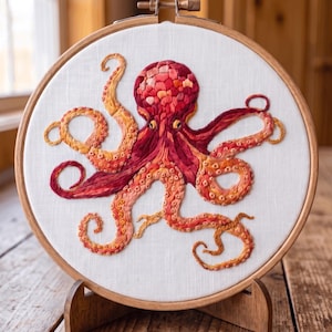 Puede incluir: Un bastidor de bordado circular de madera que contiene tela blanca con un diseño de pulpo cosido a mano. El pulpo presenta un degradado de hilos rojos, naranjas y amarillos, mostrando texturas detalladas en su cabeza y ocho tentáculos.