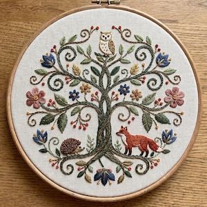 Könnte beinhalten: Ein runder Stickrahmen aus Holz mit einem weißen Stoffeinsatz, der ein handgesticktes Baummotiv zeigt. Der Baum hat einen braunen Stamm, grüne Blätter und bunte Blumen. Eine Eule sitzt oben, während ein Fuchs und ein Igel am Fuß des Baumes zu sehen sind.