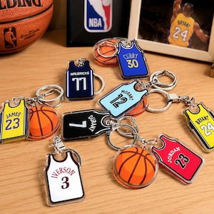 May include: A collection of basketball-themed keychains featuring miniature jerseys and basketballs. The jerseys display various player names and numbers including James 23, Curry 30, Mavericks 77, Morant 12, Durant 7, Bryant 24, Iverson 3, and Jordan 23.