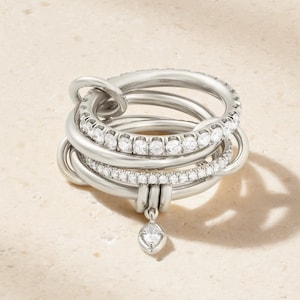 May include: A multi-band silver ring featuring several interlocking smooth metal bands and two bands encrusted with small clear stones. A single marquise-cut stone charm hangs from the bottom of the ring structure, adding a delicate detail.