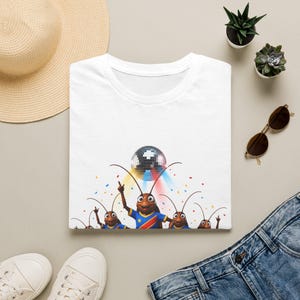 May include: A white t-shirt featuring a graphic of several brown cartoon cockroaches dancing under a disco ball. The central cockroach wears a blue shirt with a yellow star and red stripe, while colourful confetti dots are scattered around them.