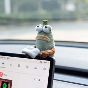 May include: A plush toy character with a sage green body, large white eyes, and a tan mouth, holding a small white cloth. The toy is perched on the top edge of a black digital car dashboard screen displaying a music interface with a Spotify logo.