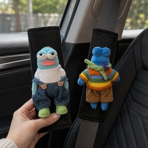 以下が含まれることがあります： 黒い布製のシートベルトカバー2点。それぞれにぬいぐるみのキャラクターが付いています。1つは水色のキャラクターで、デニムのオーバーオールと白いシャツを着ています。もう1つは青いキャラクターで、マルチカラーのストライプ柄のセーターと茶色のズボンを着用しています。