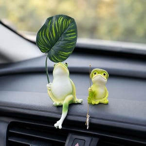 May include: Two light green frog figurines. The larger frog sits with one leg extended, holding a large, dark green, heart-shaped leaf upright. The smaller frog sits beside it, wearing a green hat and holding a thin gold fishing line with a small fish.