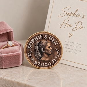 May include: A round bronze-toned commemorative coin featuring a profile portrait of a person with a ponytail, inscribed with "SOPHIE'S HEN" and "07.07.2026". Beside it sits a gold ring in a pink velvet box and a card reading "Sophie's Hen Do".