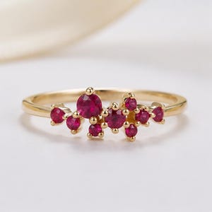 May include: A gold ring featuring a cluster of small, round, vibrant red gemstones arranged in an asymmetrical pattern across the top of the band. The metal band is polished and smooth, providing a bright contrast to the deep red stones.