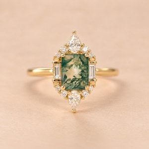 May include: A gold ring featuring a rectangular moss agate center stone with green inclusions. The stone is surrounded by a halo of small round and baguette-cut white diamonds, with a pear-shaped diamond at the top and bottom of the setting.