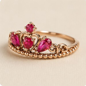 May include: A rose gold-toned ring featuring a crown-shaped design. The band is adorned with a beaded texture and set with multiple vibrant pink, heart-shaped, and round-cut gemstones arranged in a symmetrical, tiara-like formation.