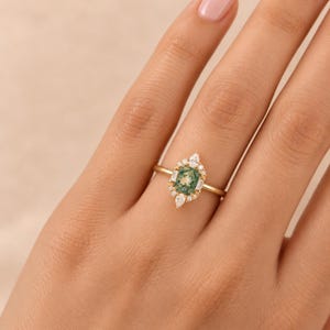 May include: A gold ring featuring a rectangular moss agate centre stone surrounded by a cluster of small, clear, faceted accent diamonds in various shapes, including pear, baguette, and round cuts, set on a thin, polished gold band.