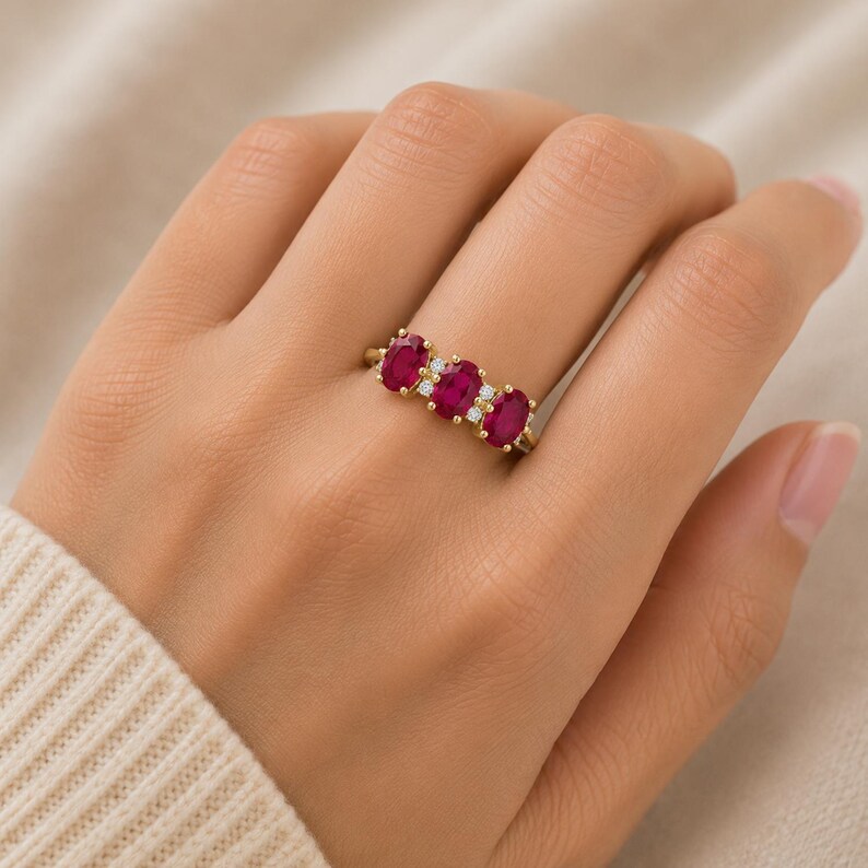Puede incluir: Un anillo de oro con tres rub&iacute;es de color rojo intenso en talla oval dispuestos horizontalmente, separados por peque&ntilde;os diamantes redondos transparentes engastados en una fina banda de oro pulido que rodea el dedo.
