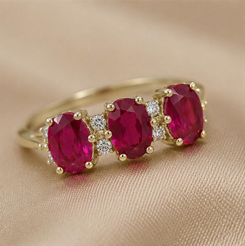 Puede incluir: Un anillo de oro con tres rub&iacute;es de color rojo intenso en talla oval dispuestos horizontalmente. Los rub&iacute;es est&aacute;n engastados en garras de oro y acentuados con peque&ntilde;os diamantes blancos redondos y claros entre cada rub&iacute;, creando un dise&ntilde;o brillante.