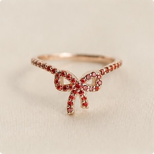 May include: A rose gold ring featuring a central bow-shaped design encrusted with small, vibrant red gemstones. The band of the ring is also adorned with a continuous row of matching red stones, creating a cohesive and detailed appearance throughout.