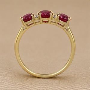 Puede incluir: Un anillo de oro con tres rub&iacute;es redondos de color rojo intenso dispuestos en fila, separados por dos peque&ntilde;as piedras de acento redondas y transparentes. El aro es liso y pulido, con cada gema sujeta firmemente por engastes de cuatro garras.