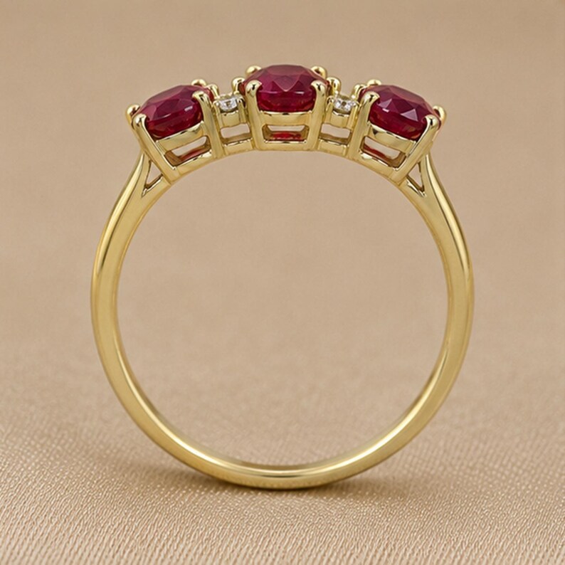 Puede incluir: Un anillo de oro con tres rub&iacute;es redondos de color rojo intenso dispuestos en fila, separados por dos peque&ntilde;as piedras de acento redondas y transparentes. El aro es liso y pulido, con cada gema sujeta firmemente por engastes de cuatro garras.