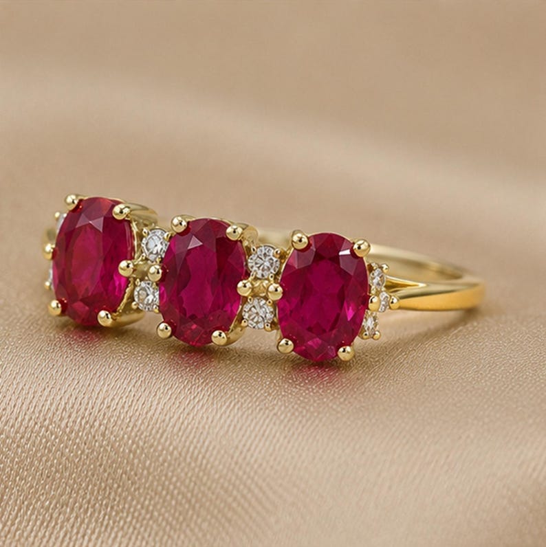 Puede incluir: Un anillo de oro con tres rub&iacute;es de color rojo intenso en talla oval dispuestos horizontalmente. Cada rub&iacute; est&aacute; rodeado por peque&ntilde;as piedras de acento transparentes de talla redonda engastadas en oro. El aro es de oro amarillo pulido.