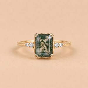 May include: A gold ring featuring a rectangular emerald-cut moss agate center stone with dark green dendritic inclusions. The band is accented with two small round white stones on each side and has a textured, beaded metal setting.
