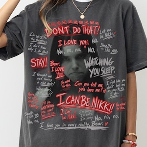 May include: A charcoal grey oversized t-shirt featuring a central black and white portrait of a person surrounded by red and white handwritten-style text. The graphic includes phrases like DONT DO THAT, I LOVE YOU, and I CAN BE NIKKI.