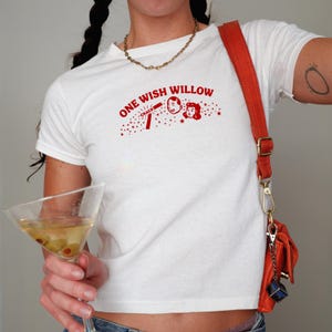 May include: A white short-sleeved t-shirt featuring a red graphic print on the chest. The design includes the text "ONE WISH WILLOW" above a cartoon illustration of two faces, a "CRACK" sound effect, and scattered small stars and dots.