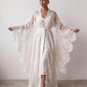 May include: A floor-length white lace robe featuring wide, flared sleeves with an intricate fan-like pattern and a scalloped edge. The garment includes a matching white fabric tie belt at the waist and is worn over a simple white slip dress.