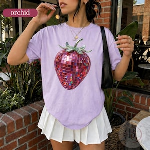 May include: A light purple short-sleeve t-shirt featuring a large graphic of a strawberry made of reflective, multi-colored disco ball tiles with green leaves on top. The shirt has a crew neckline and a loose, relaxed fit.