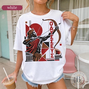 May include: A white t-shirt featuring a graphic print of a bearded king wearing a crown and holding a bow and arrow. The design includes red and black geometric shapes, a Greek column, a small ship, and the Greek text OAYΣΣEYΣ in vertical red letters.
