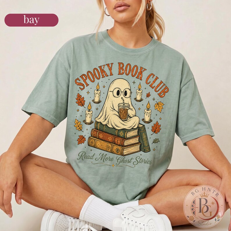 Pode incluir: Uma t-shirt de manga curta verde-s&aacute;lvia com um gr&aacute;fico de um fantasma de &oacute;culos, segurando uma bebida e sentado numa pilha de livros. O texto diz "SPOOKY BOOK CLUB" e "Read More Ghost Stories" com ilustra&ccedil;&otilde;es de velas e folhas de outono.