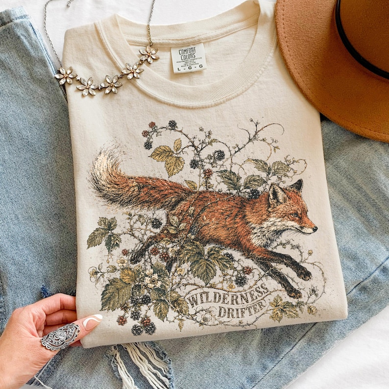 May include: A cream-colored t-shirt featuring a graphic of a fox running through blackberry vines and leaves. The text "WILDERNESS DRIFTER" is printed in an arch below the fox. The shirt has a Comfort Colors label indicating size L-G.