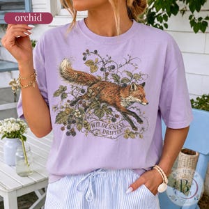 May include: A light purple t-shirt featuring a graphic illustration of a red fox leaping through blackberry vines and leaves. The text "WILDERNESS DRIFTER" is printed in a decorative font below the fox. The shirt has a crew neckline and short sleeves.