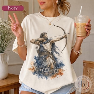 May include: An ivory t-shirt featuring a watercolour-style graphic of a bearded archer aiming a bow and arrow. The figure wears a draped tunic and is surrounded by blue and brown paint splatters. Below the figure, the Greek text OΔYΣΣEYΣ is printed.