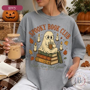Pode incluir: Uma t-shirt cinzenta com um gr&aacute;fico de um fantasma de &oacute;culos, sentado numa pilha de livros enquanto segura uma bebida. O texto diz "SPOOKY BOOK CLUB" e "Read More Ghost Stories", rodeado por velas acesas e folhas de outono.