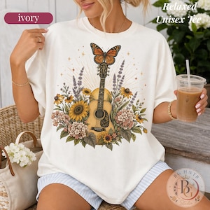 May include: An ivory short-sleeve t-shirt featuring a graphic print of an acoustic guitar surrounded by sunflowers, lavender, and a monarch butterfly. The design includes radiating sunburst lines behind the guitar. The text reads Relaxed Unisex Tee.