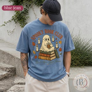 Pode incluir: Uma t-shirt azul jeans com um gr&aacute;fico de um fantasma de &oacute;culos, sentado numa pilha de livros enquanto segura uma bebida. O texto "SPOOKY BOOK CLUB" e "Read More Ghost Stories" est&aacute; rodeado por velas acesas e folhas de outono.