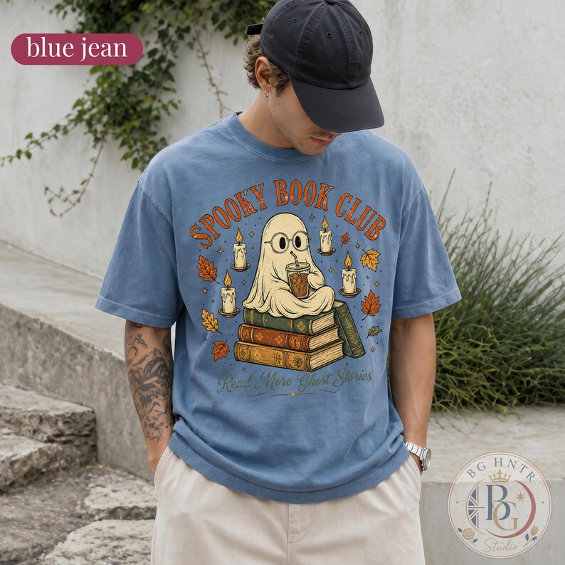 Pode incluir: Uma t-shirt azul jeans com um gr&aacute;fico de um fantasma de &oacute;culos, sentado numa pilha de livros enquanto segura uma bebida. O texto "SPOOKY BOOK CLUB" e "Read More Ghost Stories" est&aacute; rodeado por velas acesas e folhas de outono.