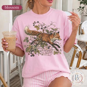 May include: A light pink t-shirt featuring a graphic illustration of a fox running through blackberry vines. The text "WILDERNESS DRIFTER" is printed in a curved banner below the fox. The shirt has a crew neckline and short sleeves.