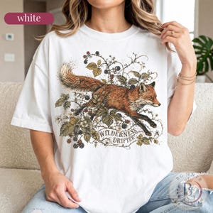 May include: A white short-sleeved t-shirt featuring a detailed graphic of a fox leaping through blackberry vines. The design includes the text "WILDERNESS DRIFTER" in a curved banner below the fox, rendered in earthy tones of brown, green, and black.