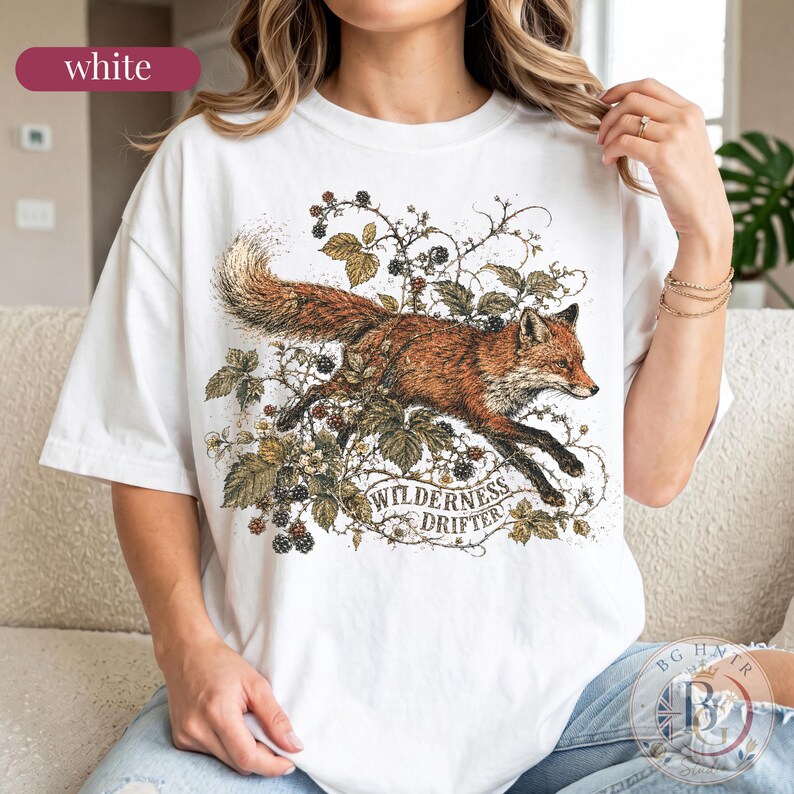 May include: A white short-sleeved t-shirt featuring a detailed graphic of a fox leaping through blackberry vines. The design includes the text "WILDERNESS DRIFTER" in a curved banner below the fox, rendered in earthy tones of brown, green, and black.