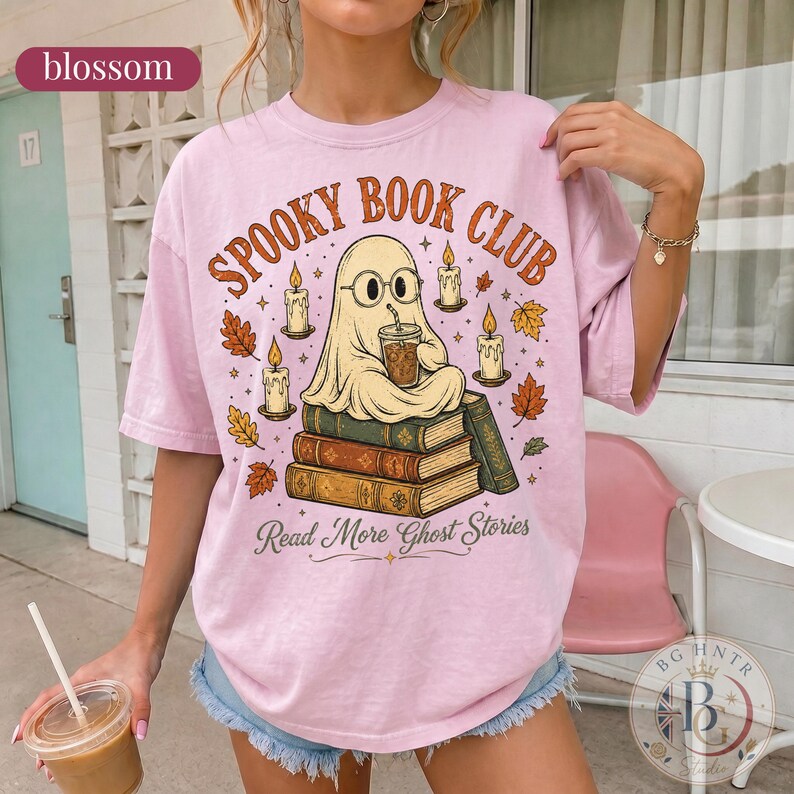 Pode incluir: Uma t-shirt rosa claro com um gr&aacute;fico de um fantasma de &oacute;culos, sentado numa pilha de livros enquanto segura uma bebida. O texto diz SPOOKY BOOK CLUB e Read More Ghost Stories, rodeado por ilustra&ccedil;&otilde;es de velas e folhas de outono.