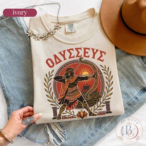 May include: An ivory t-shirt featuring a graphic of a Greek archer aiming a bow, with the Greek text "ΟΔΥΣΣΕΥΣ" arched above. The design includes a sun, a ship on waves, and laurel branches. The shirt has a Comfort Colors label at the neck.