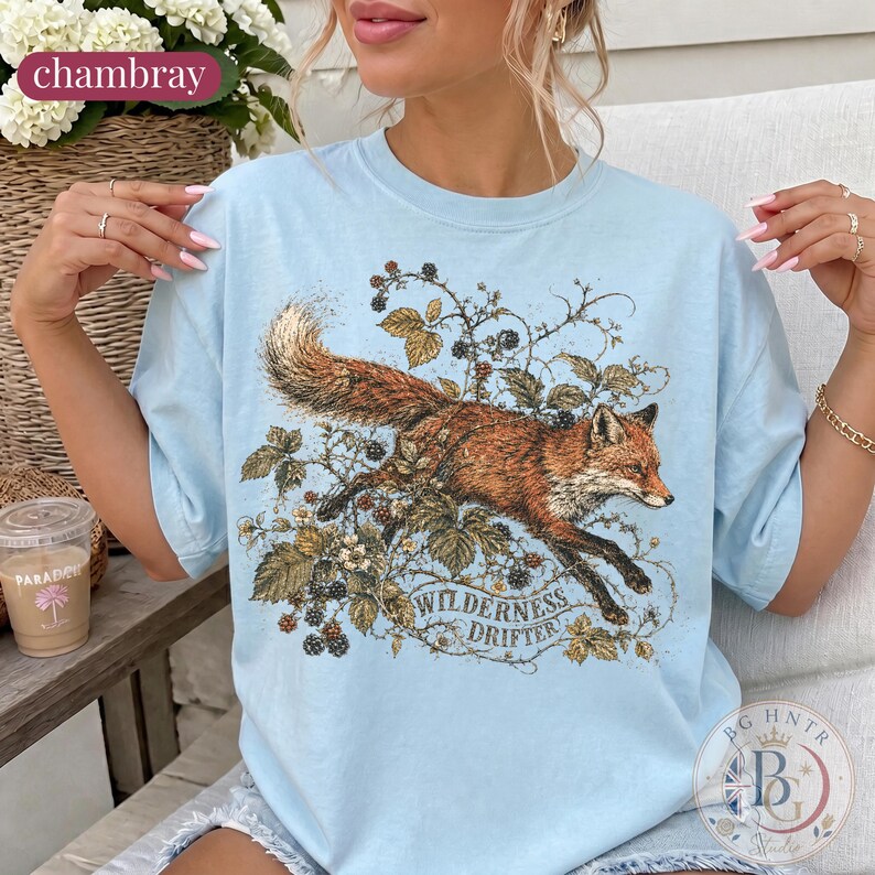 May include: A light blue t-shirt featuring a graphic illustration of a red fox leaping through blackberry vines and leaves. The text "WILDERNESS DRIFTER" is printed in a decorative font below the fox. The shirt has a crew neckline and short sleeves.