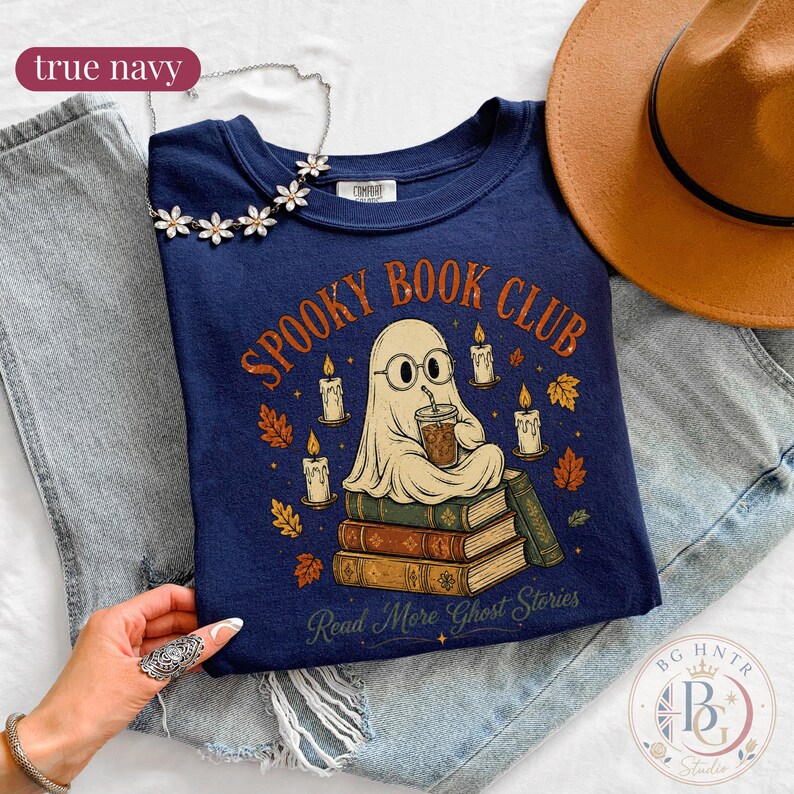 Pode incluir: Uma t-shirt azul-marinho com um gr&aacute;fico de um fantasma de &oacute;culos, segurando uma ch&aacute;vena de caf&eacute; e sentado numa pilha de livros. O texto diz "SPOOKY BOOK CLUB" e "Read More Ghost Stories" rodeado por velas e folhas de outono.