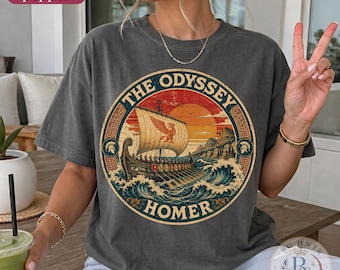 Puede incluir: Una camiseta gris carbón con un estampado gráfico circular de un antiguo barco griego navegando sobre olas hacia una puesta de sol. El texto dice THE ODYSSEY y HOMER. El diseño incluye un emblema alado en la vela y un borde de greca griega.