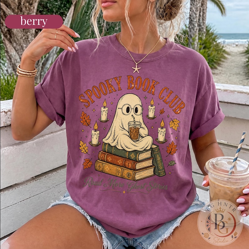 Pode incluir: Uma t-shirt de manga curta cor de baga com um gr&aacute;fico de um fantasma de &oacute;culos sentado numa pilha de livros enquanto bebe de um copo. O texto "SPOOKY BOOK CLUB" est&aacute; escrito em letras cor de laranja, rodeado por velas e folhas de outono.