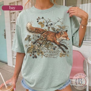 May include: A sage green short-sleeve t-shirt featuring a detailed graphic of a red fox leaping through blackberry vines and leaves. The text "WILDERNESS DRIFTER" is printed in a curved font below the fox illustration on the front of the shirt.
