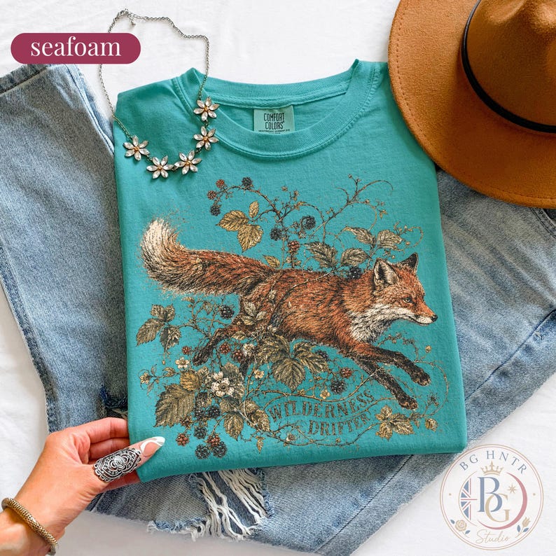 May include: A seafoam green t-shirt featuring a graphic illustration of a fox running through blackberry vines. The text "WILDERNESS DRIFTER" is printed below the fox. The shirt has a Comfort Colors label at the neck and is paired with blue jeans.