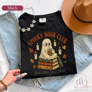 Pode incluir: Uma t-shirt preta com um gr&aacute;fico de um fantasma de &oacute;culos segurando um copo de caf&eacute; enquanto est&aacute; sentado numa pilha de livros. O texto diz "SPOOKY BOOK CLUB" e "Read More Ghost Stories", rodeado por velas e folhas de outono.