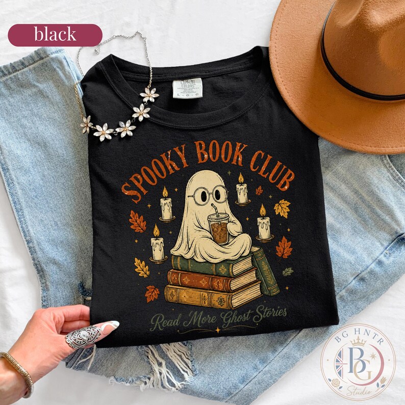 Pode incluir: Uma t-shirt preta com um gr&aacute;fico de um fantasma de &oacute;culos segurando um copo de caf&eacute; enquanto est&aacute; sentado numa pilha de livros. O texto diz "SPOOKY BOOK CLUB" e "Read More Ghost Stories", rodeado por velas e folhas de outono.