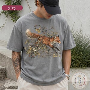 May include: A grey short-sleeve t-shirt featuring a graphic illustration of a fox leaping through blackberry vines and leaves. The text "WILDERNESS DRIFTER" is printed in a subtle font beneath the fox. The shirt has a crew neckline and a relaxed fit.