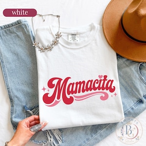 May include: A white short-sleeve t-shirt featuring the word Mamacita in a red and pink cursive font with small star and heart accents. The shirt is paired with blue denim jeans, a brown wide-brimmed hat, and a silver-toned flower necklace.