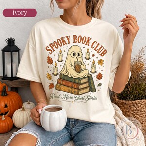 Pode incluir: Uma t-shirt cor de marfim com um gr&aacute;fico de um fantasma de &oacute;culos, sentado numa pilha de livros enquanto segura uma bebida. O texto diz "SPOOKY BOOK CLUB" e "Read More Ghost Stories", rodeado por ilustra&ccedil;&otilde;es de velas acesas e folhas de outono.