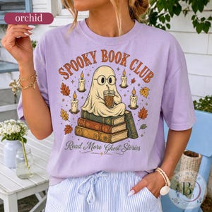 Pode incluir: Uma t-shirt roxo-claro com um gr&aacute;fico de um fantasma de &oacute;culos, segurando uma ch&aacute;vena de caf&eacute; e sentado numa pilha de livros. O texto diz SPOOKY BOOK CLUB e Read More Ghost Stories, rodeado por ilustra&ccedil;&otilde;es de velas e folhas de outono.